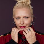 Portrait of a blonde woman with her hair in a bun, wearing a red jacket with a stand-up collar and black inserts, with a visible set of silver rings on her fingers and decorative earrings and studs in her ear.