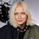 Portrait of a blonde woman with medium-length hair and subtle makeup, wearing a dark jacket and a decorative, expressive necklace with large, round beads against the background of a light wall with an abstract pattern.