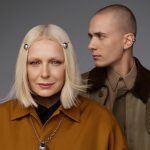 Portrait of two models against a grey background – in the foreground a blonde woman with decorative ball-shaped hair clips in her hair and a necklace, and in the background a shaved man with an earring, dressed in brown and beige clothing.