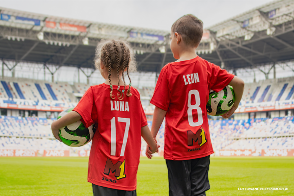Dwoje dzieci trzyma się za ręce na stadionie piłkarskim, trzymają piłki. Zdjęcie pokazuje dzieci od tyłu, w czerwonych koszulkach piłkarskich - dziewczynka po lewej stronie ma koszulkę z numerem 17 i imieniem Lena, chłopiec stojący po prawej z numerem 8 i imieniem Leon.