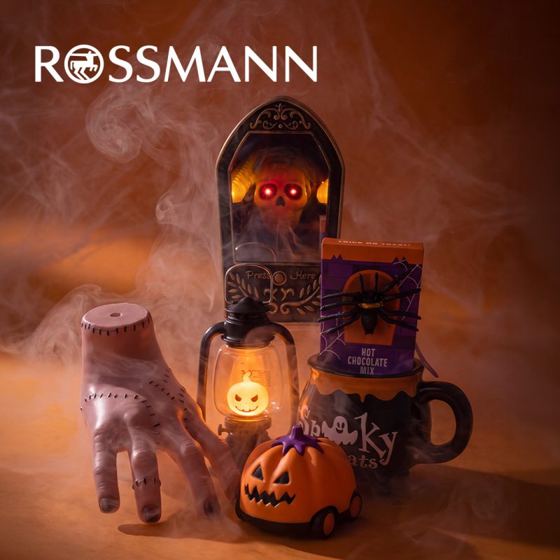 Dekoracje na Halloween na pomarańczowym tle spowitym mgłą: sztuczna ręka, dzwonek z czaszką o świecących czerwonych oczach, latarnia z podświetlaną dynią, pojazd w kształcie dyni oraz kubek z czekoladą do picia. U góry białe logo Rossmann.